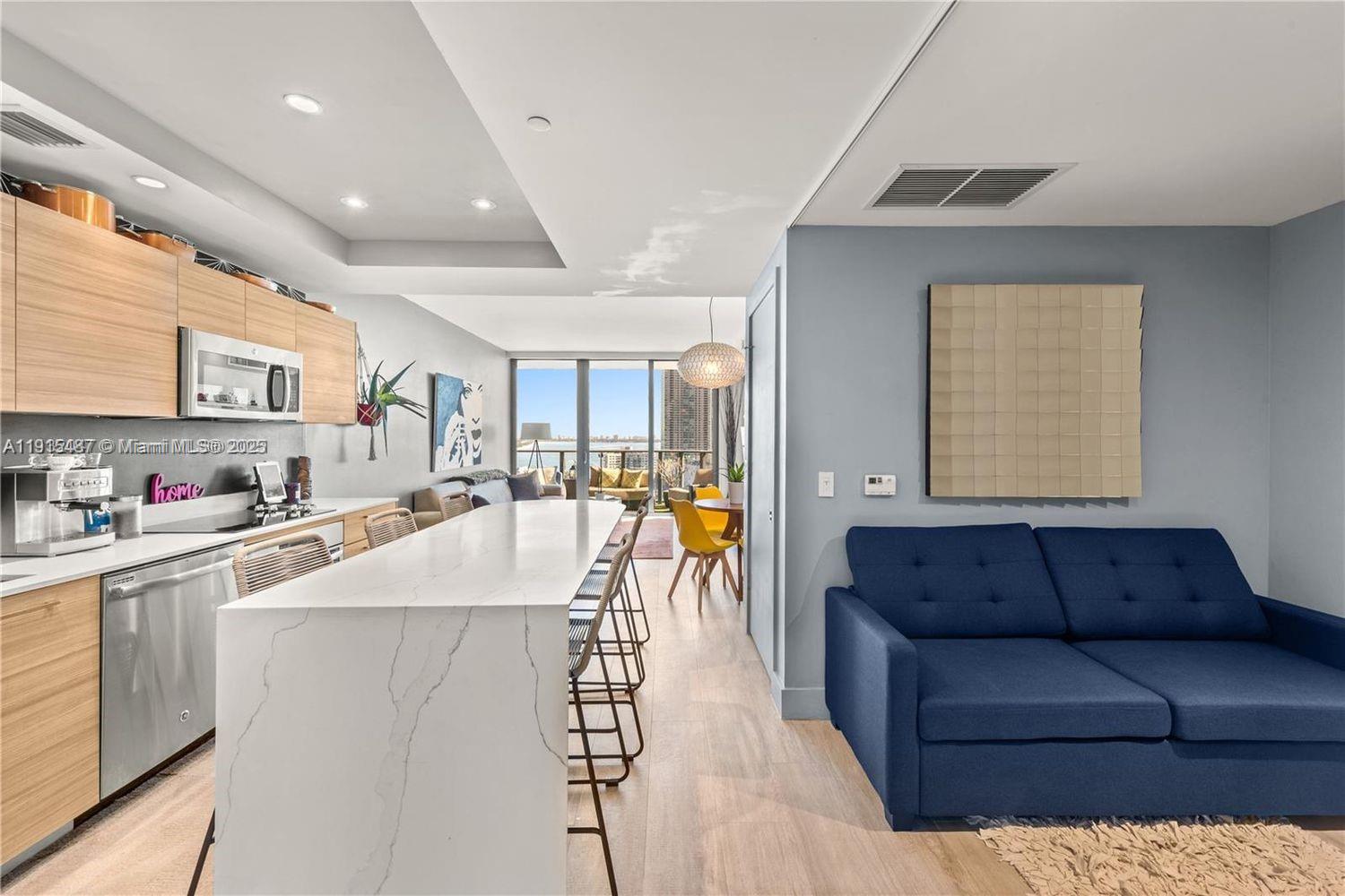 An open-concept kitchen and living area features a white island, blue sofa, and balcony view — 121 NE 34th St # 1705, Miami FL 33137, MIAMI, FL