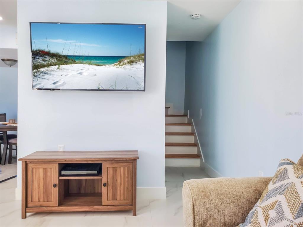 A living room features a wall-mounted television above a wooden media console and stairs — 215 MEDALLION BOULEVARD UNIT G, MADEIRA BEACH, FL