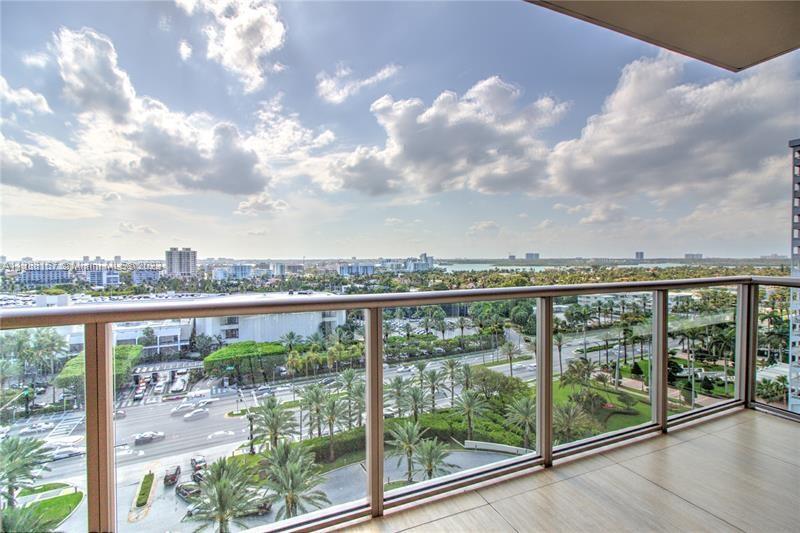 A balcony with a glass railing overlooks a city street, palm trees, and distant water — 9705 Collins Ave # 1003N, Bal Harbour FL 33154, BAL HARBOUR, FL