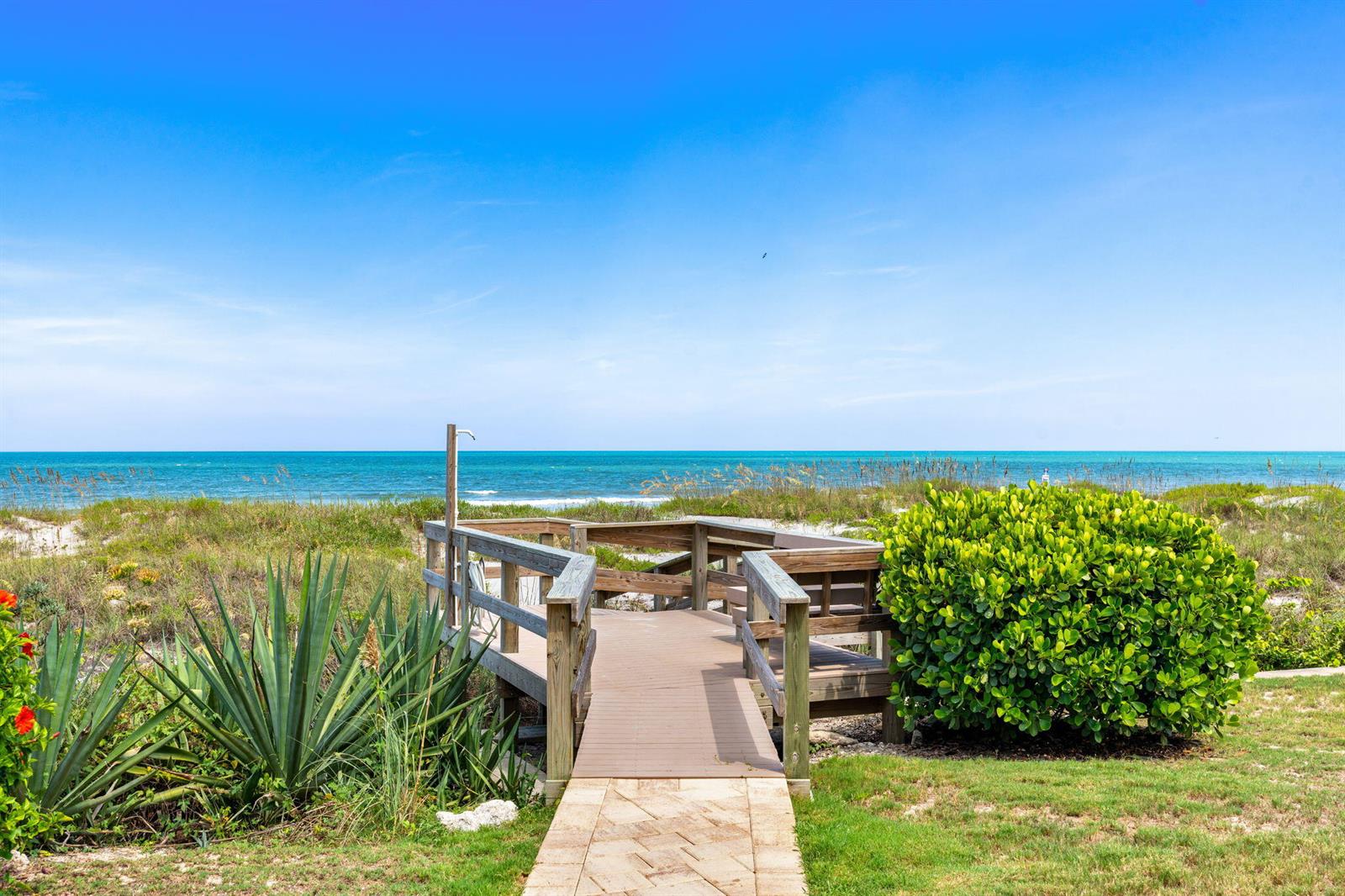 A wooden boardwalk leads through coastal vegetation toward the ocean under a clear blue sky — 1155 S Atlantic Avenue # 203, Cocoa Beach FL 32931, COCOA BEACH, FL
