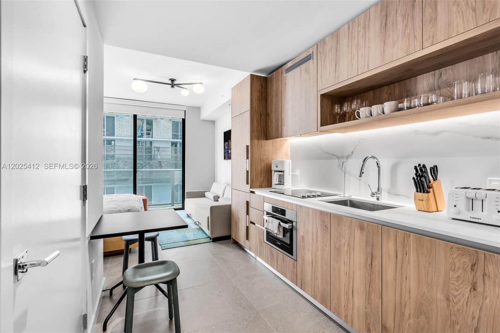 A modern kitchen with wood-toned cabinetry and a small dining area overlooks a living space — 11 NE 6th St # 2608, Miami FL 33132, MIAMI, FL