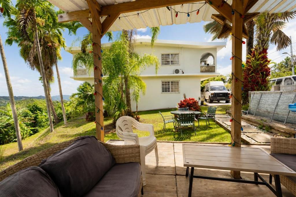 A covered patio with outdoor furniture overlooks a grassy yard and a two-story house — Carr 156 KM 1.0 BO SABANA SECTOR PUEBLITO, OROCOVIS, PR
