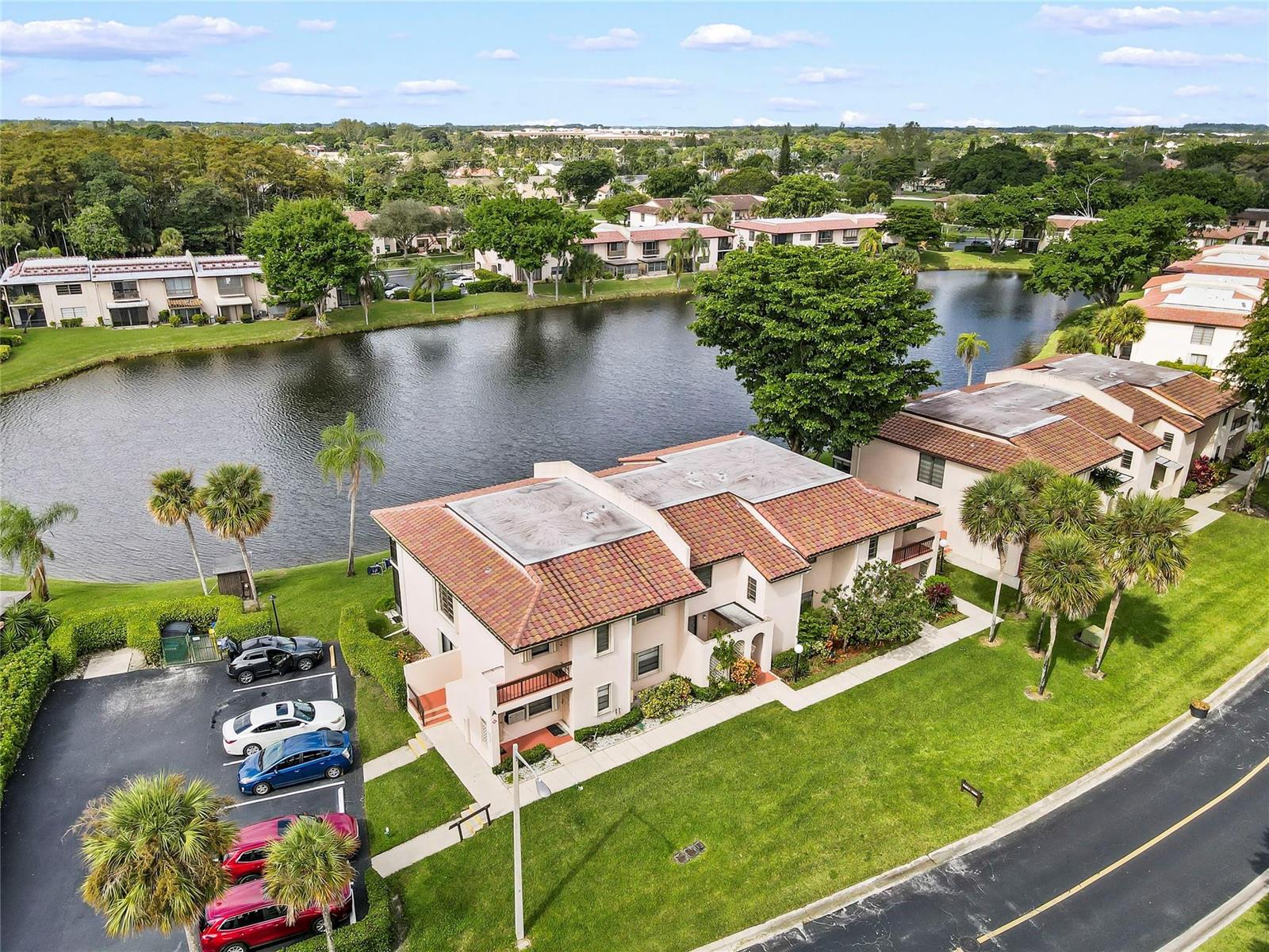 An aerial view shows multi-story apartment buildings with tile roofs situated beside a lake — 21649 Juego Circle, Boca Raton FL 33433, BOCA RATON, FL