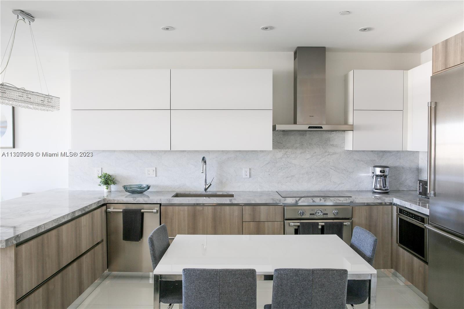 A modern kitchen features white upper cabinets, wood-tone lower cabinets, marble countertops, and a dining table — 100 Lincoln Rd # 1643, Miami Beach FL 33139, MIAMI BEACH, FL