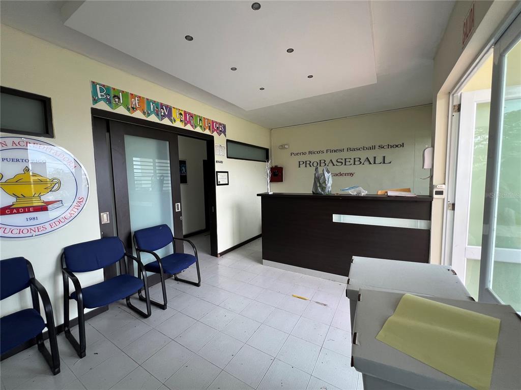 A reception area features a dark wood desk, blue chairs, and a baseball academy sign — Marginal PR-52 BARRIO MONTELLANOS, SECTOR PEPE HOYOS, CAYEY, PR