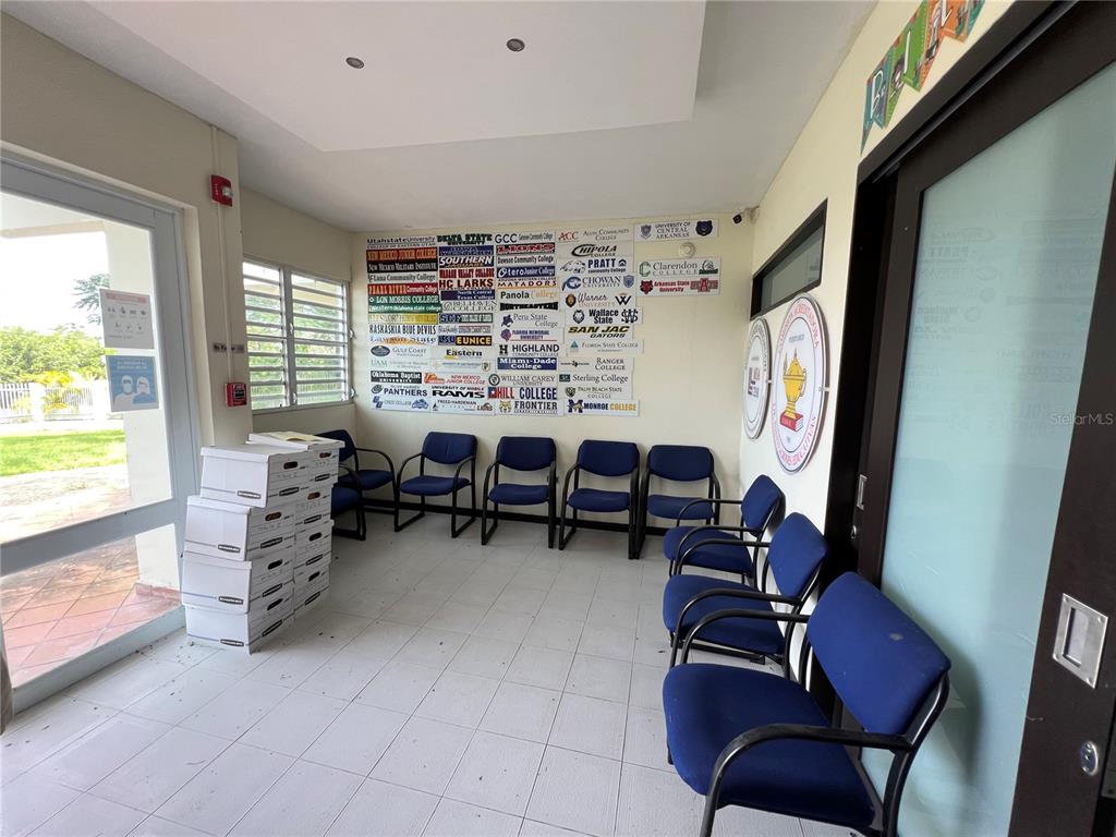 A waiting room features blue chairs, a tiled floor, and a wall covered in college logos — Marginal PR-52 BARRIO MONTELLANOS, SECTOR PEPE HOYOS, CAYEY, PR
