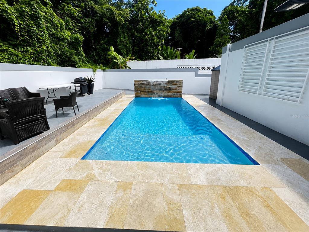 A rectangular swimming pool with a stone water feature sits on a light-colored stone patio — . ESMERALDA UNIT E-12, SAN JUAN, PR