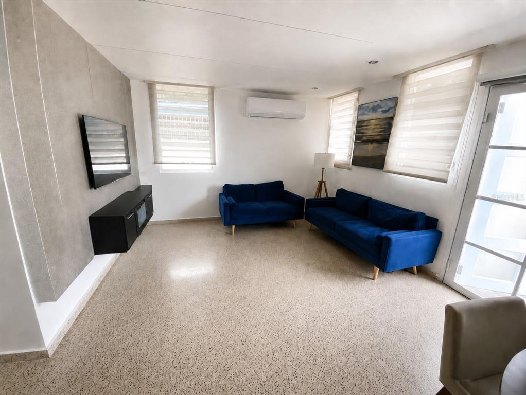 A living room features blue sofas, a wall-mounted television, and light-colored terrazzo flooring — . ESMERALDA UNIT E-12, SAN JUAN, PR
