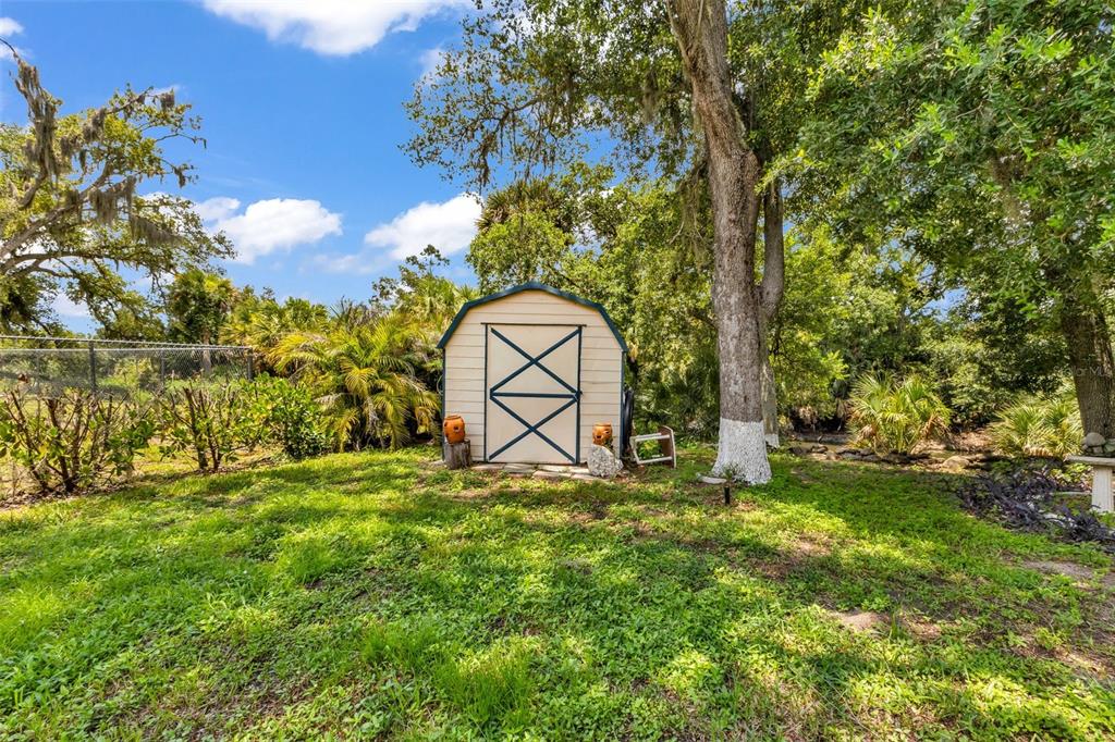 A tan storage shed with blue trim sits in a grassy yard under large trees — 297 FLAMINGO BOULEVARD, PORT CHARLOTTE, FL
