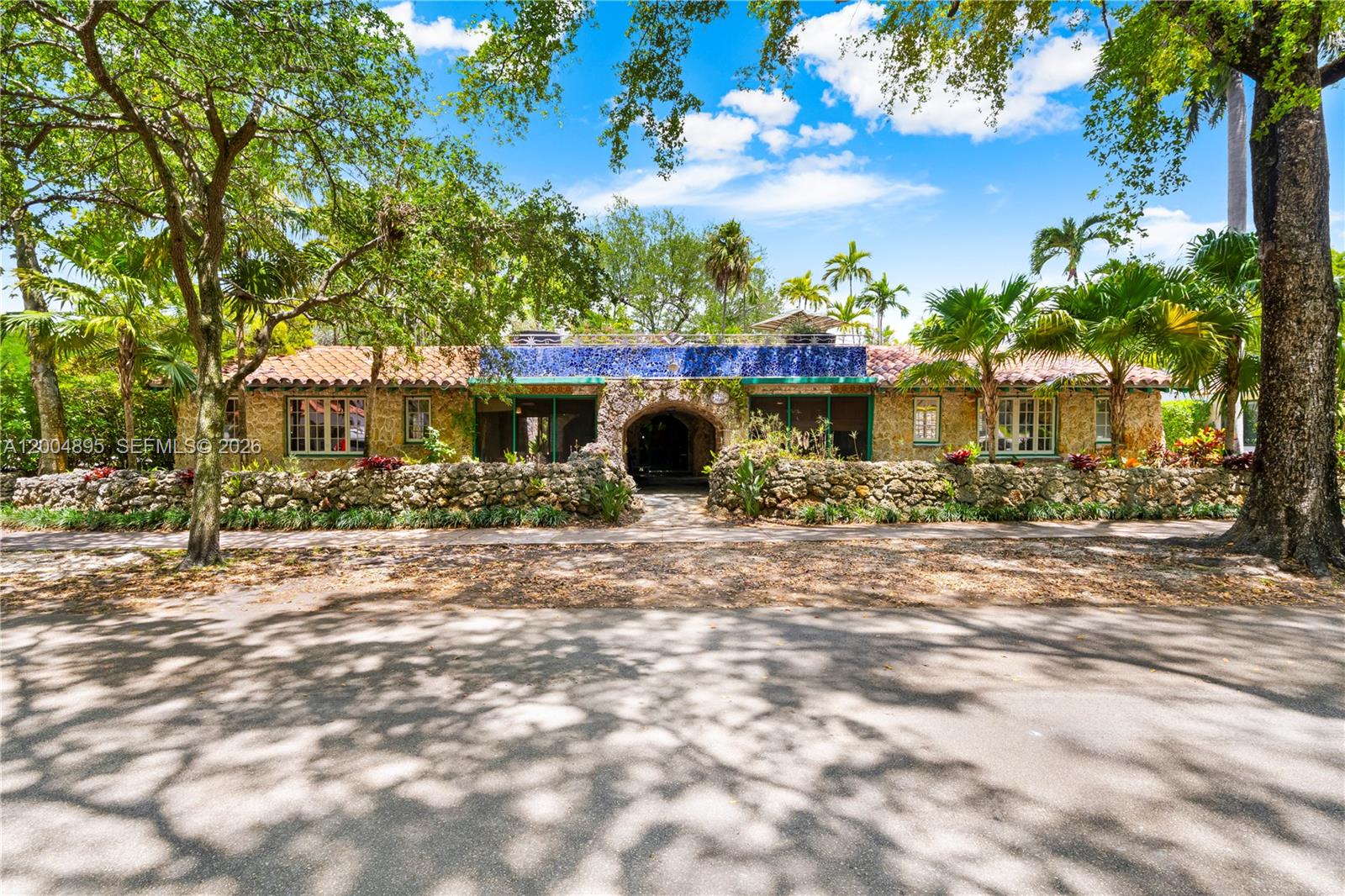 A single-story stone house with a blue tiled facade and arched entryway sits behind trees — 1301 Milan Ave, Coral Gables FL 33134, CORAL GABLES, FL