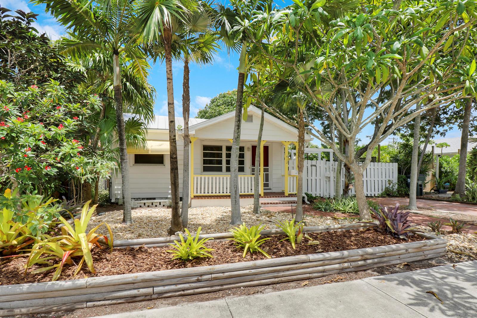 A white cottage with a yellow-trimmed porch sits behind a landscaped garden with palm trees — 412 N K Street, FL 33460, FL