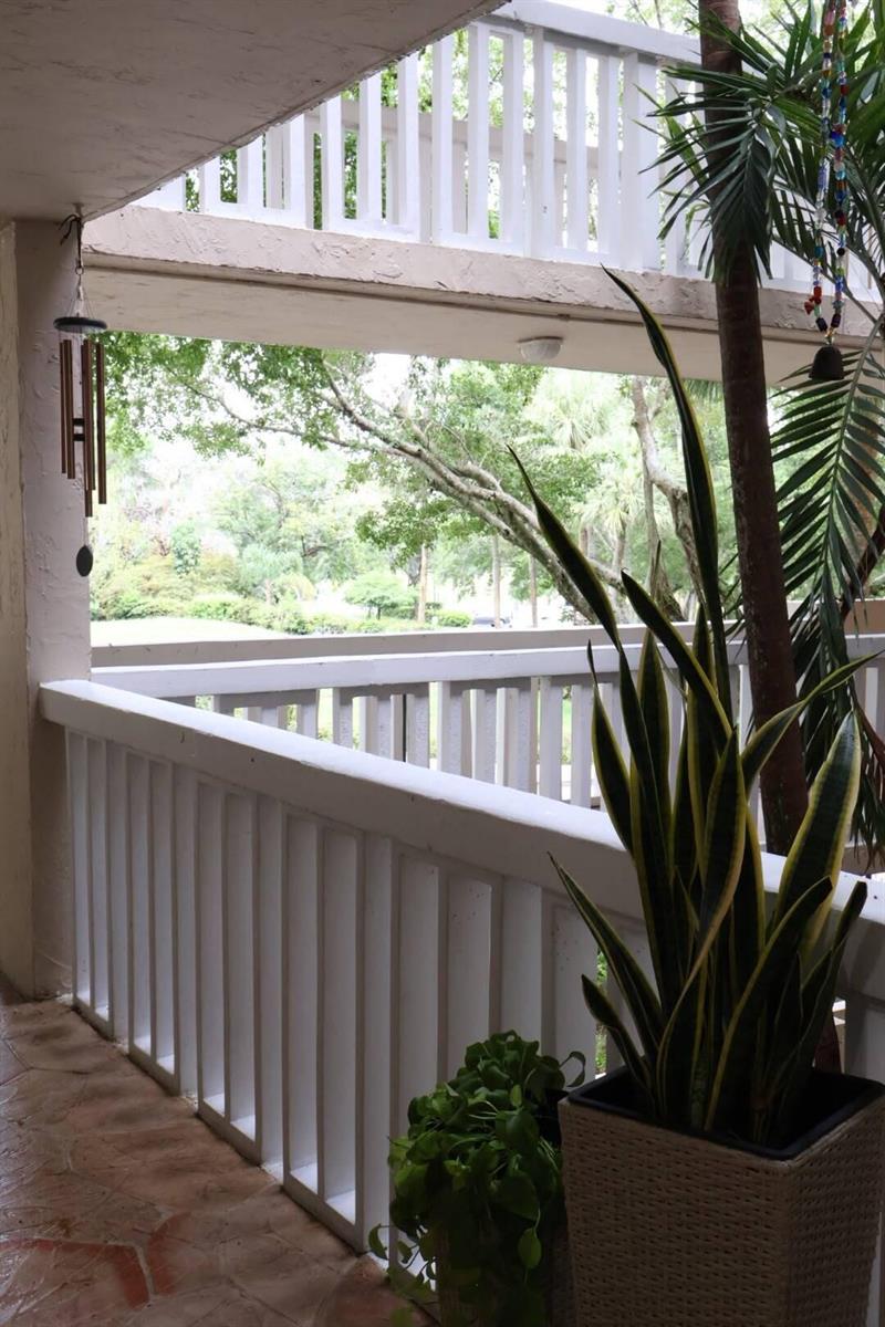 A covered balcony features white railings, a potted snake plant, and a view of trees — 7011 Environ Boulevard # 210, Lauderhill FL 33319, LAUDERHILL, FL