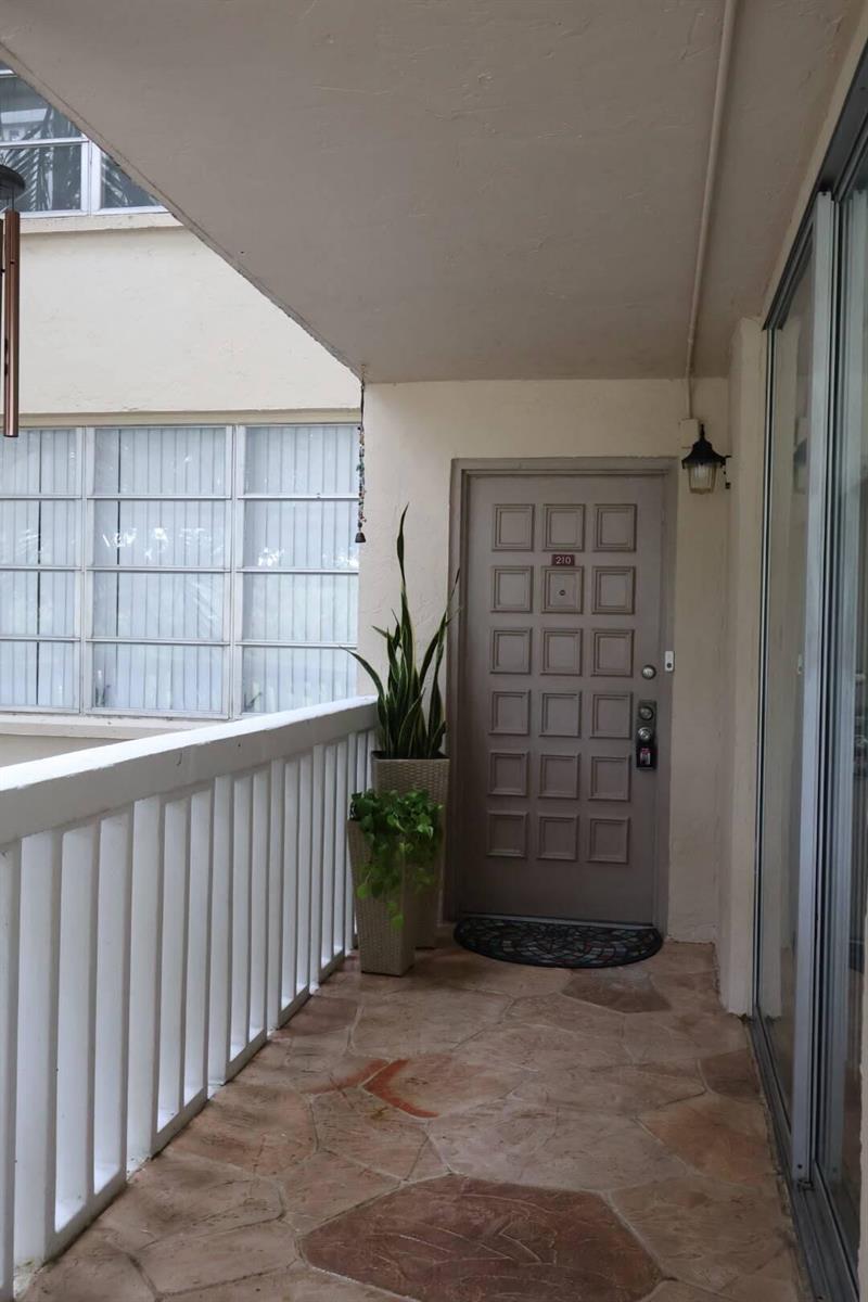 An outdoor apartment entryway features a gray paneled door, stone flooring, and potted plants — 7011 Environ Boulevard # 210, Lauderhill FL 33319, LAUDERHILL, FL