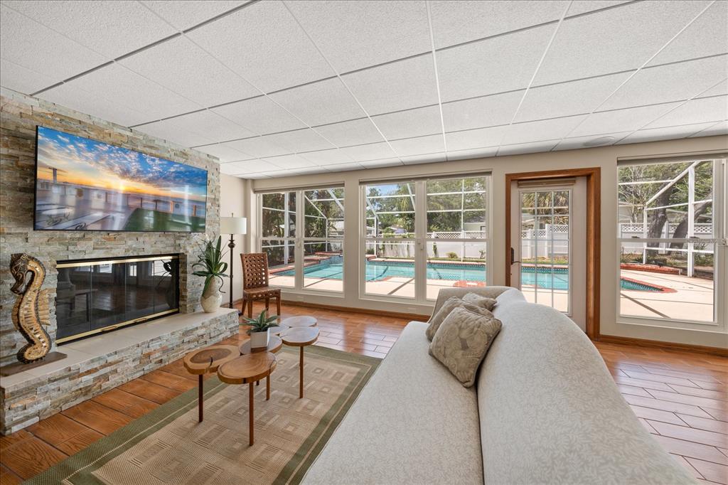 A living room with a stone fireplace, wood-look tile floors, and views of a pool — 802 ALLAMANDA DRIVE, LARGO, FL