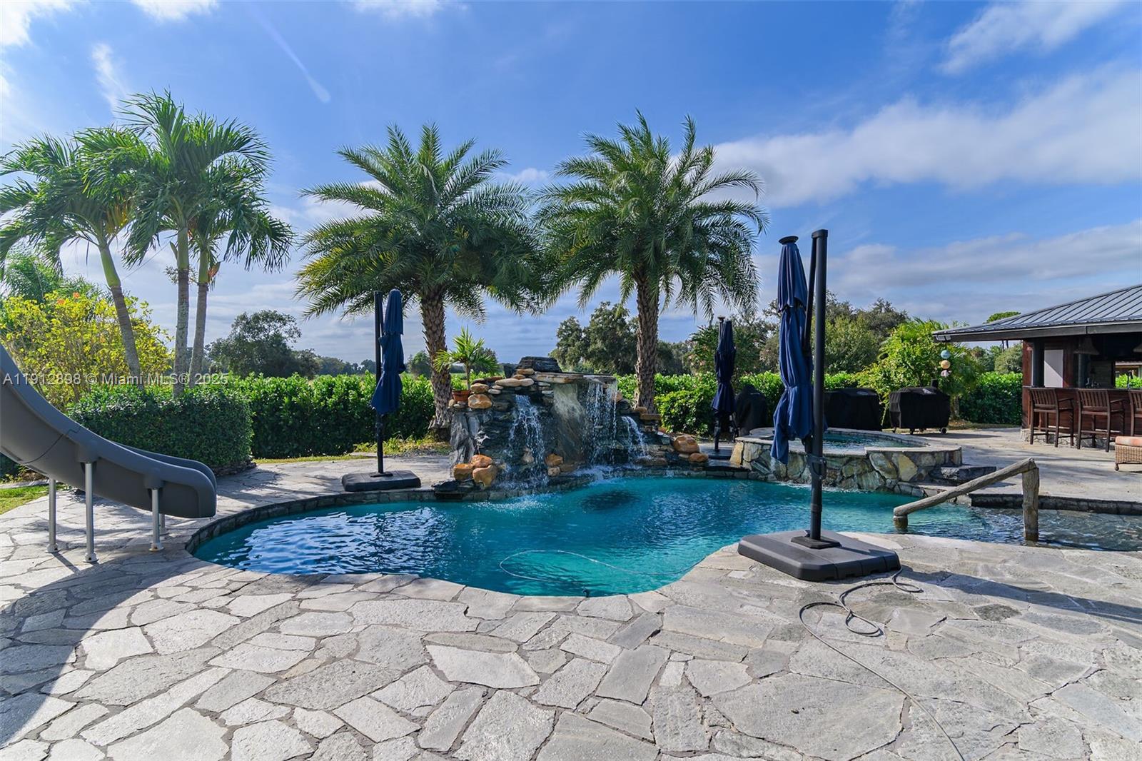 A backyard swimming pool features a rock waterfall, a slide, and stone patio flooring — 5675 County Road 78, La Belle FL 33935, LA BELLE, FL