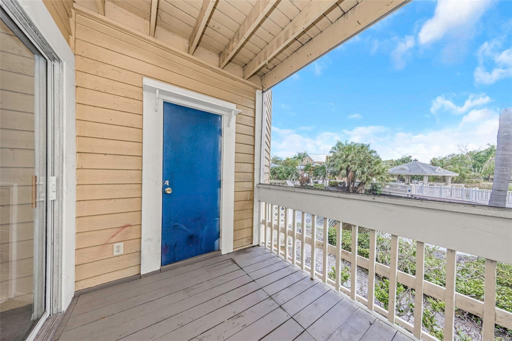 A wooden balcony features a blue door, light-colored siding, and a white railing overlooking greenery — 115 112TH AVENUE NE UNIT 703, ST PETERSBURG, FL