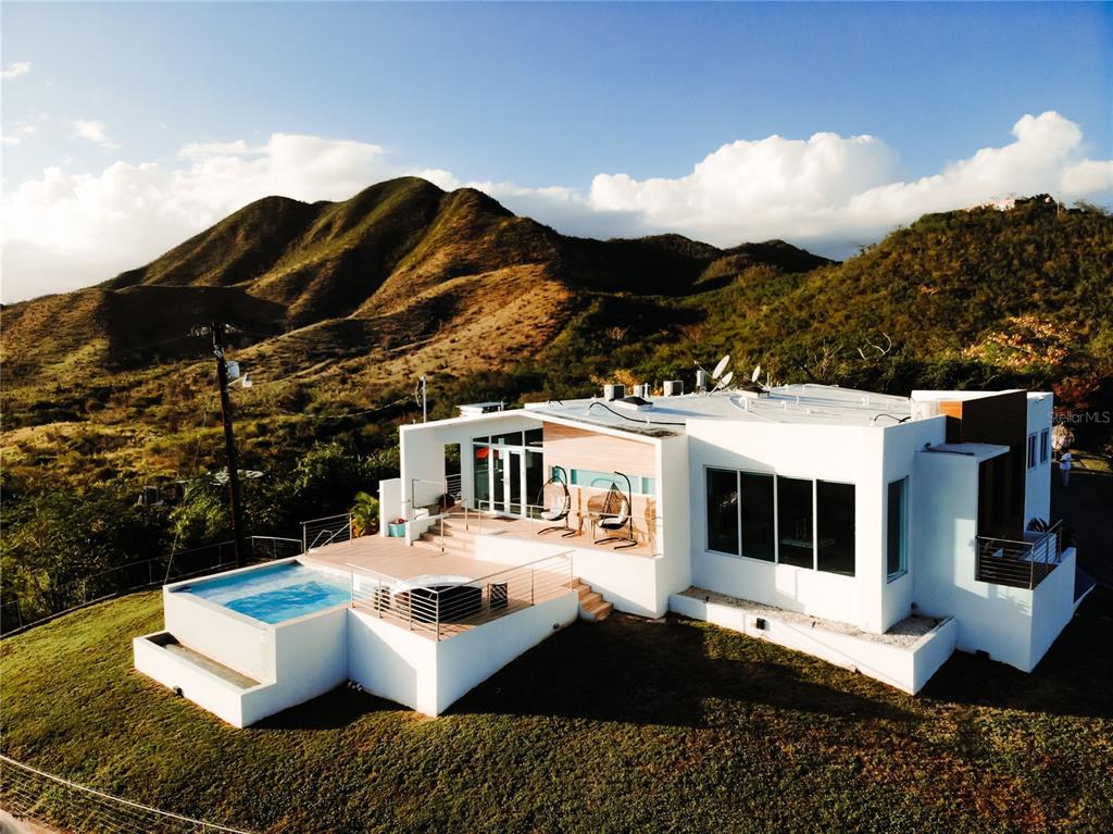 An aerial view shows a modern white house with wood accents nestled in hilly terrain — KM 0.7 PR-551, JUANA DIAZ, PR