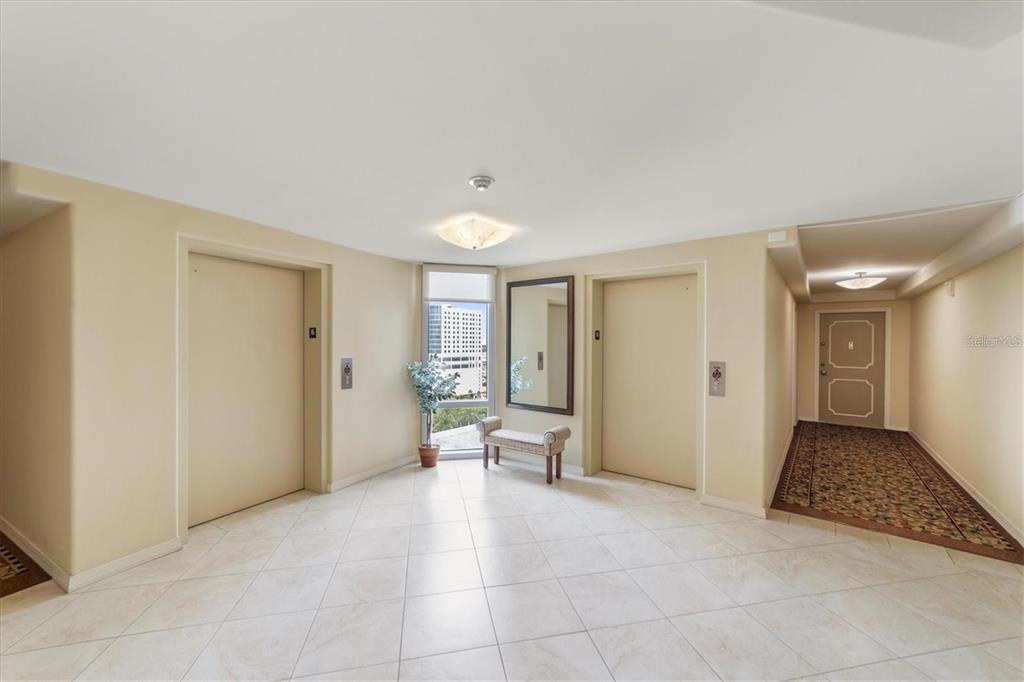 A building elevator lobby with beige walls, tile flooring, a bench, and a window view — 1111 N GULFSTREAM AVENUE UNIT 16C, SARASOTA, FL