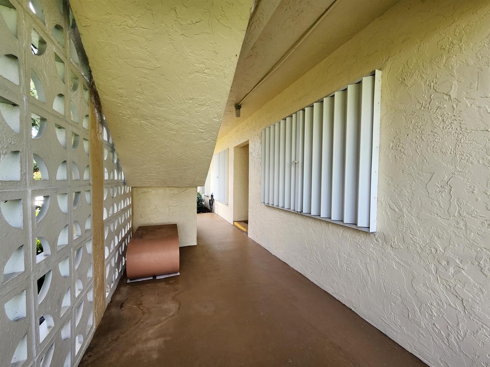 An exterior walkway features a decorative concrete block wall, beige stucco, and metal storm shutters — 1560 NW 18th Avenue # 104, Delray Beach FL 33445, DELRAY BEACH, FL