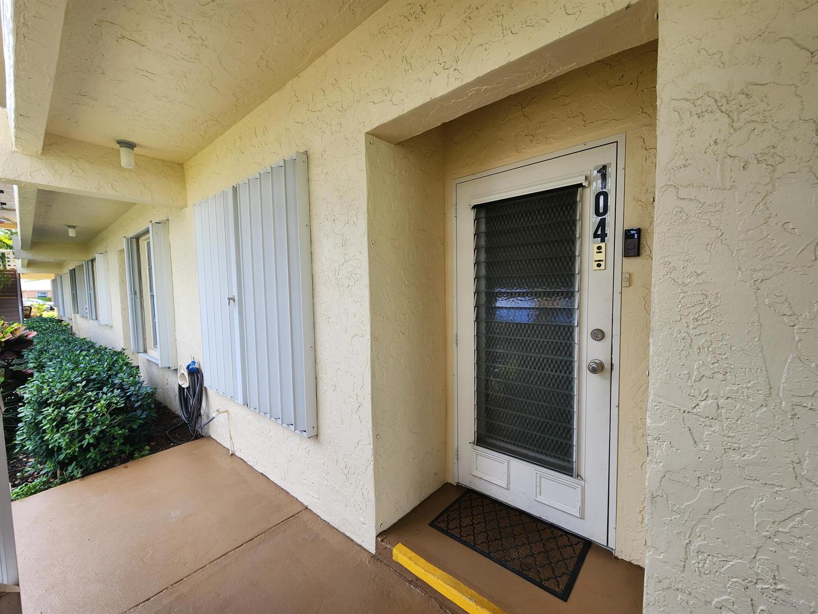 An exterior view of a residential unit entrance with a white door and metal shutters — 1560 NW 18th Avenue # 104, Delray Beach FL 33445, DELRAY BEACH, FL
