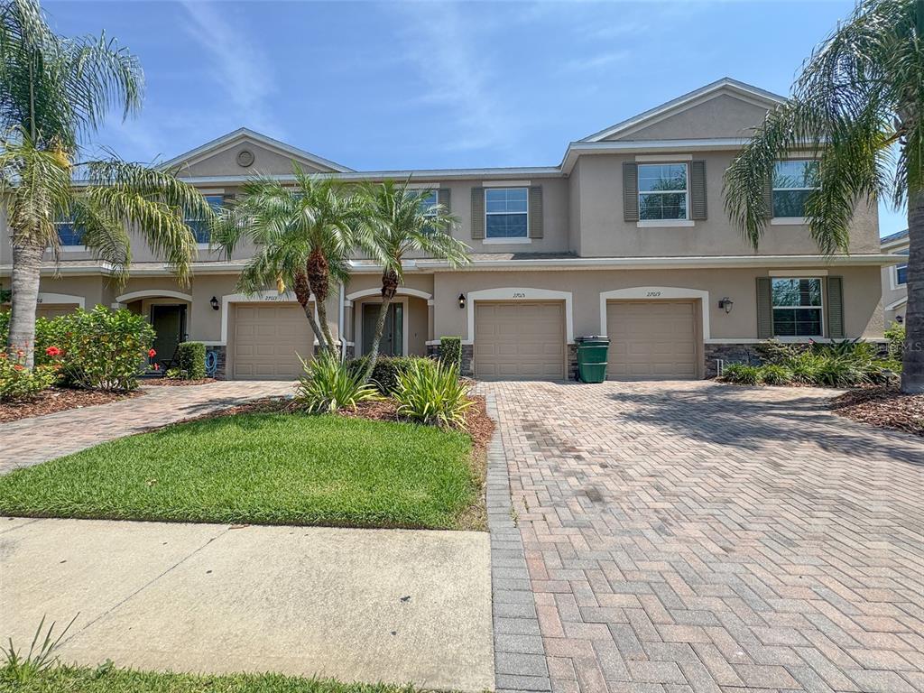 A two-story townhouse exterior features tan stucco walls, multiple garages, and a paved driveway — 27015 JUNIPER BAY DRIVE, WESLEY CHAPEL, FL
