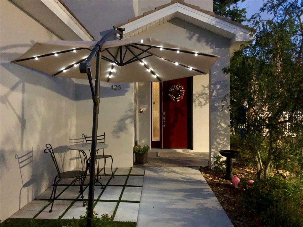 A patio with a lit umbrella and bistro set sits before a house entrance — 426 BIRDSONG LANE, DUNEDIN, FL