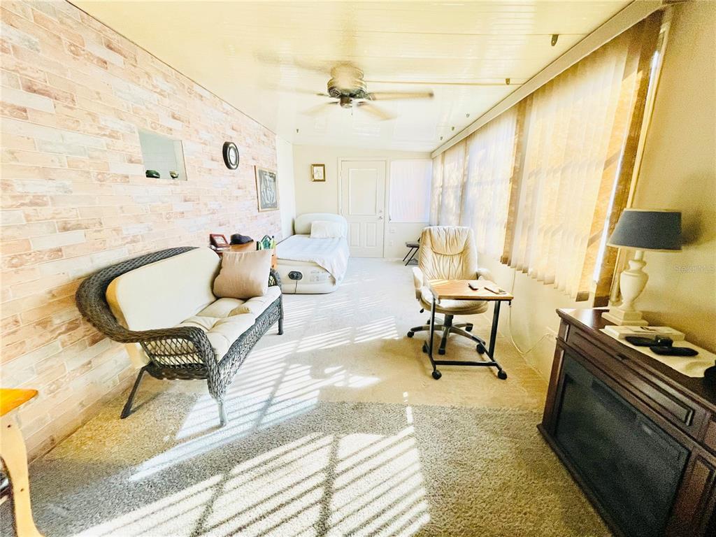 A sunroom features a brick-patterned wall, carpeted flooring, a ceiling fan, and several furniture pieces — 3275 26TH AVENUE E UNIT 107, BRADENTON, FL