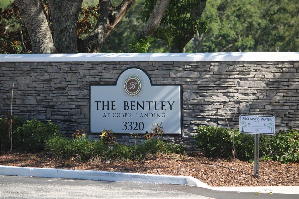 A stone wall features a sign for The Bentley at Cobb's Landing with landscaping — 3300 HAVILAND COURT UNIT 104, PALM HARBOR, FL