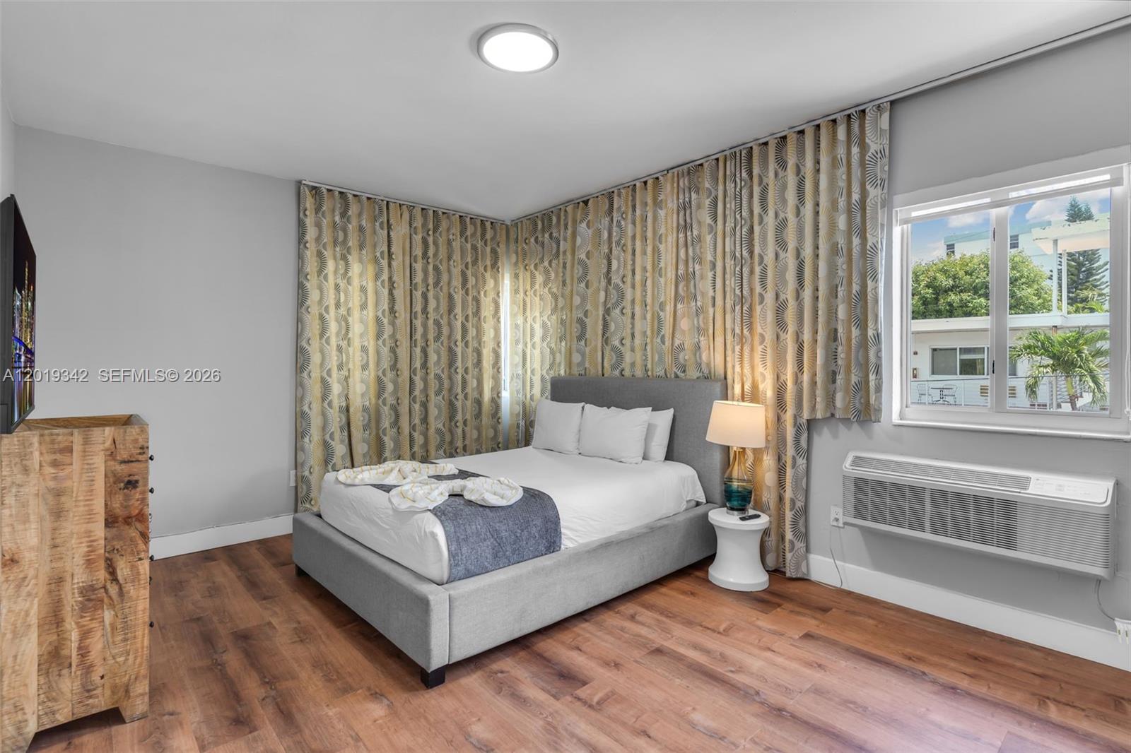 A bedroom features a gray upholstered bed, patterned curtains, wood-look flooring, and a wall-mounted air conditioner — 8100 Harding Ave # 514, Miami Beach FL 33141, MIAMI BEACH, FL