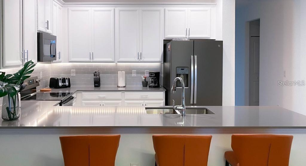 A modern kitchen features white cabinets, gray countertops, stainless steel appliances, and three orange stools — 1139 DROP LANE UNIT 1139, DAVENPORT, FL