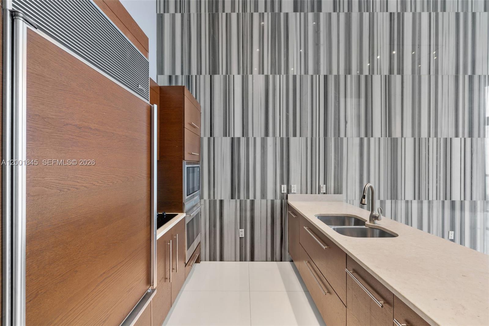 A modern kitchen features wood cabinetry, a double sink, and a tall, striped marble backsplash — 475 Brickell Ave # 2812, Miami FL 33131, MIAMI, FL