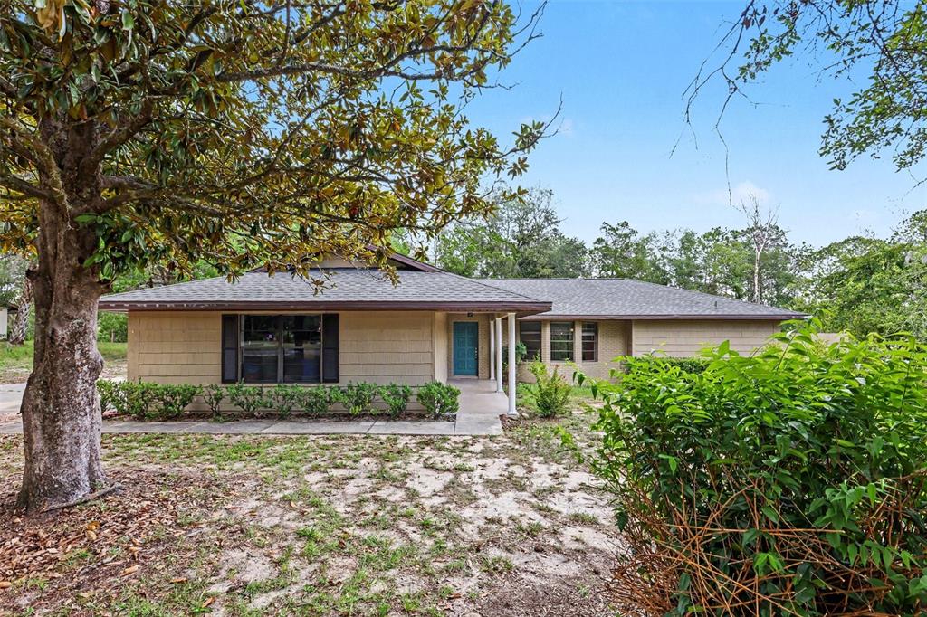 A single-story tan house with a dark roof sits behind a large tree and shrubs — 9491 SW 200TH COURT, DUNNELLON, FL