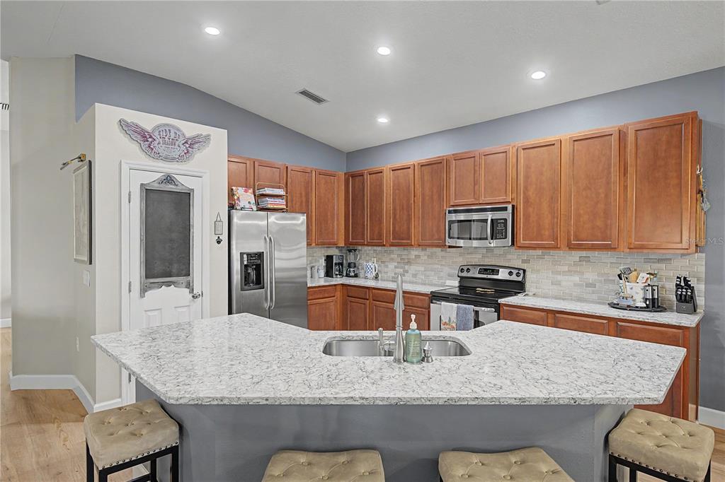 A kitchen features wood cabinets, granite countertops, a central island with seating, and stainless appliances — 34726 PINEHURST GREENE WAY, ZEPHYRHILLS, FL