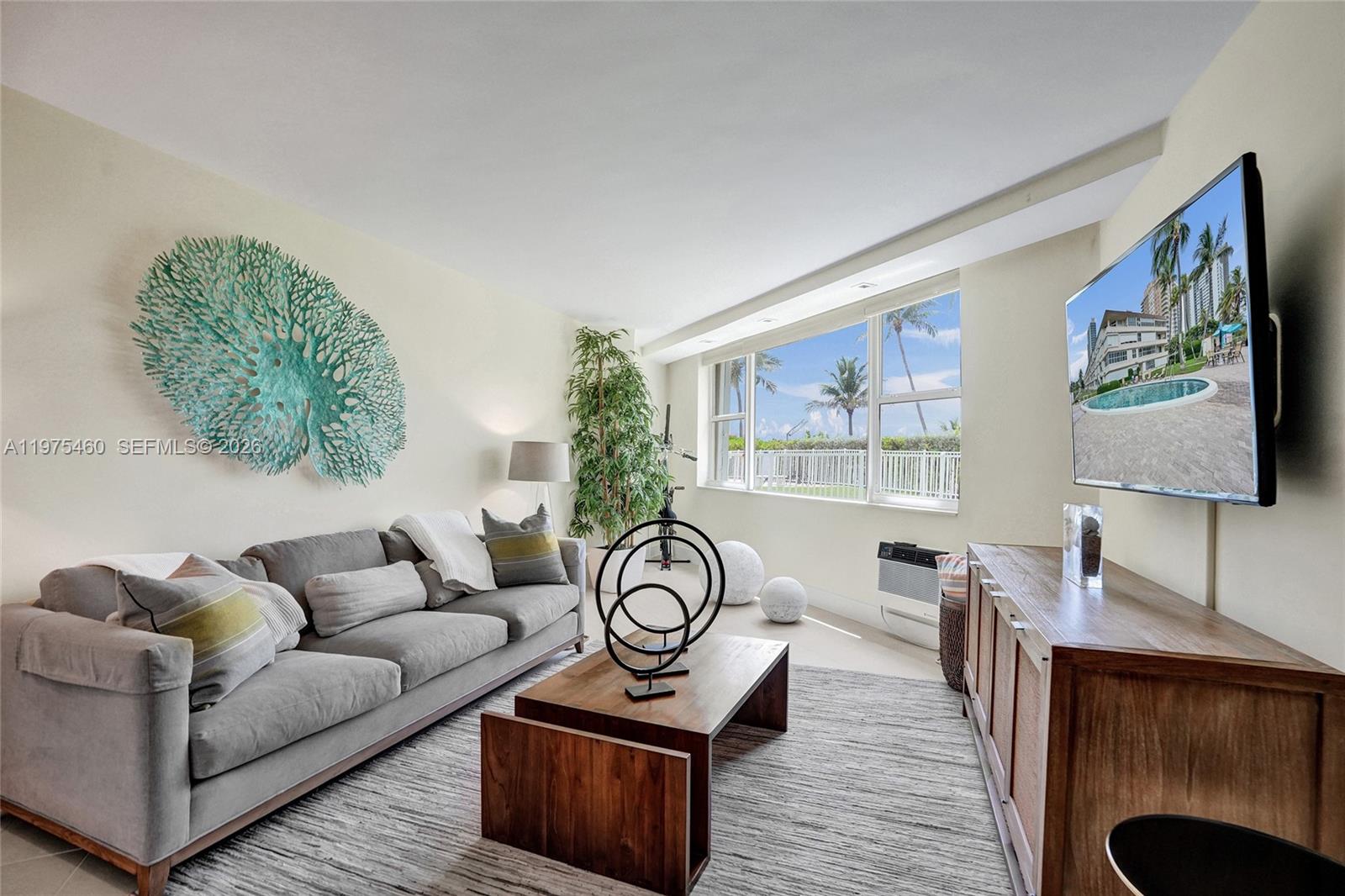 A living room features a gray sofa, wooden coffee table, wall-mounted television, and large windows — 1936 S Ocean # 21D, Hallandale Beach FL 33009, HALLANDALE BEACH, FL