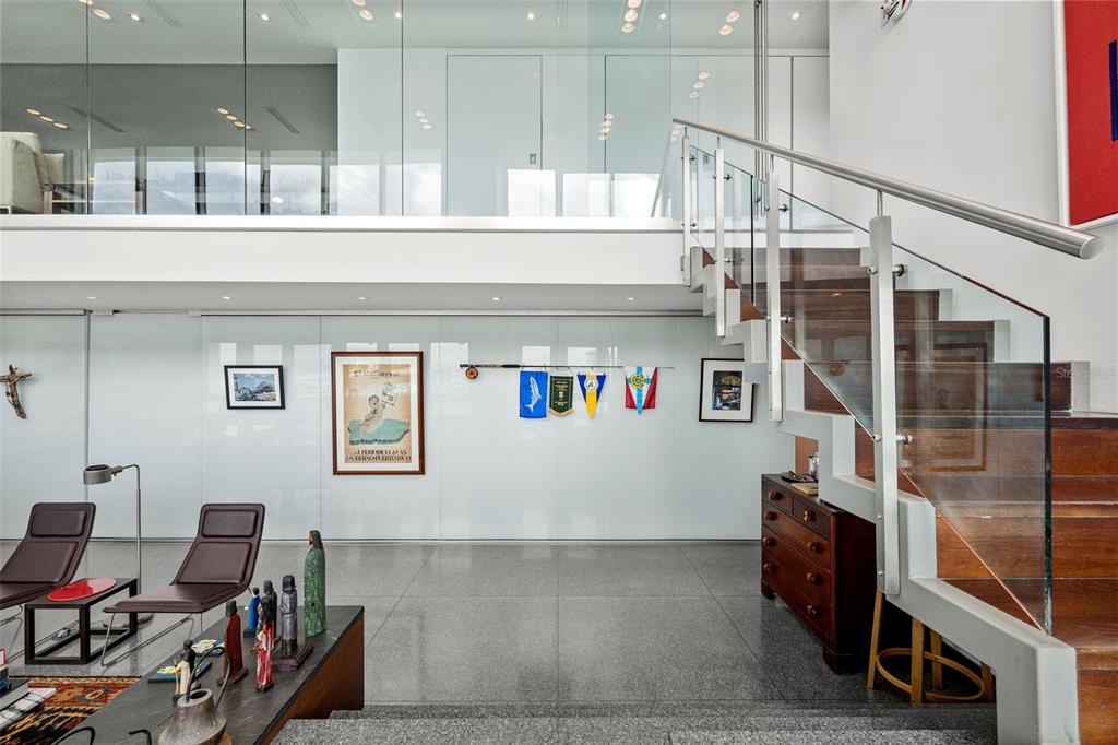 A modern living room features a glass-railed wooden staircase, polished stone floors, and white walls — 550 AVENIDA CONSTITUCION UNIT PH-1608, SAN JUAN, PR