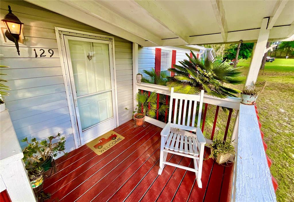 A white rocking chair sits on a red-painted wooden porch with a welcome mat — 129 CRYSTAL LAKE DRIVE, MELROSE, FL