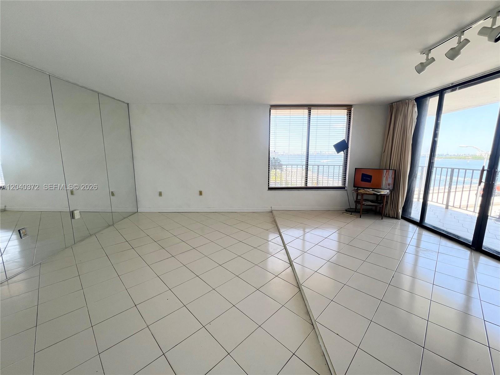 A living room features white tiled floors, mirrored walls, and a balcony with water views — 7899 NE Bayshore Ct # 6A, Miami FL 33138, MIAMI, FL