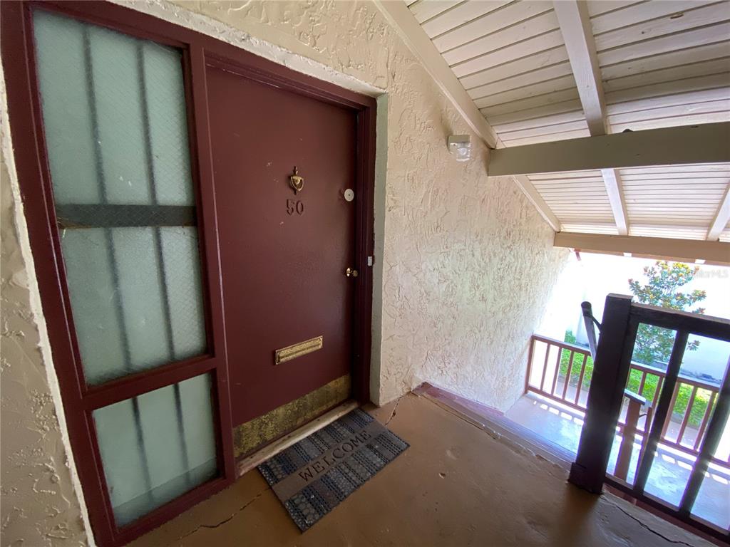 A red front door with a side window and a welcome mat on a balcony — 8602 HURON COURT UNIT 50, TAMPA, FL