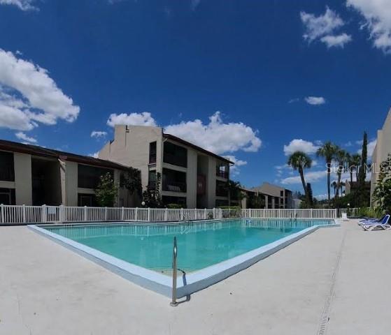 A rectangular swimming pool sits in front of multi-story apartment buildings under a blue sky — 8602 HURON COURT UNIT 50, TAMPA, FL