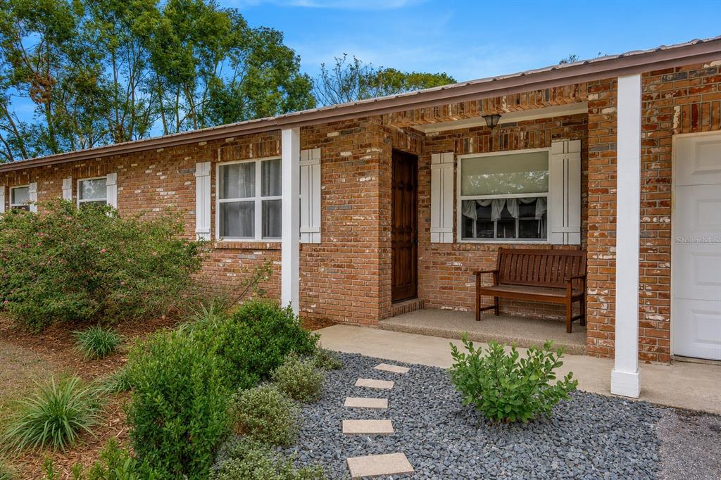 A brick house exterior features a covered porch, a wooden bench, and a gravel walkway — 12212 FLORAL LANE, THONOTOSASSA, FL