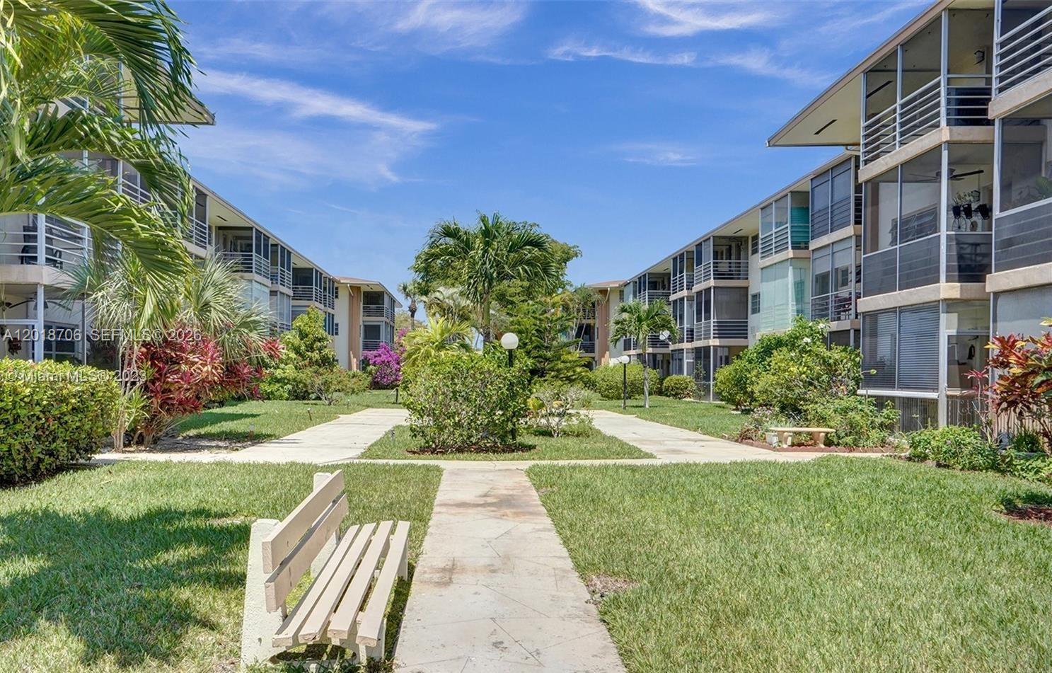 A courtyard with a concrete path, a wooden bench, and palm trees between two apartment buildings — 17090 NE 14th Ave # 312, North Miami Beach FL 33162, NORTH MIAMI BEACH, FL