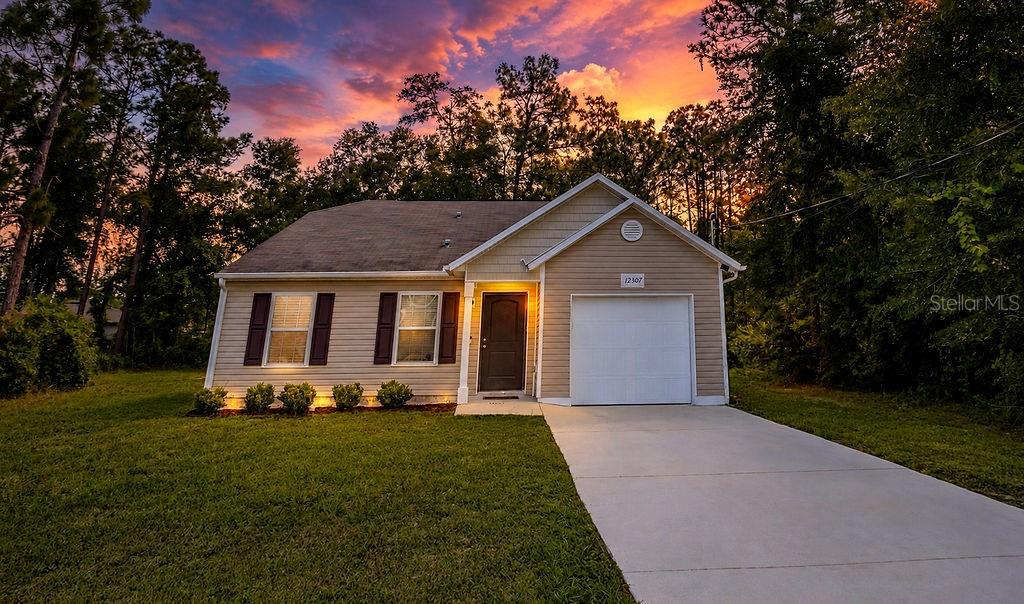 A tan, single-story house with a white garage door and a concrete driveway at sunset — 12307 MILLINGTON AVENUE, WEEKI WACHEE, FL