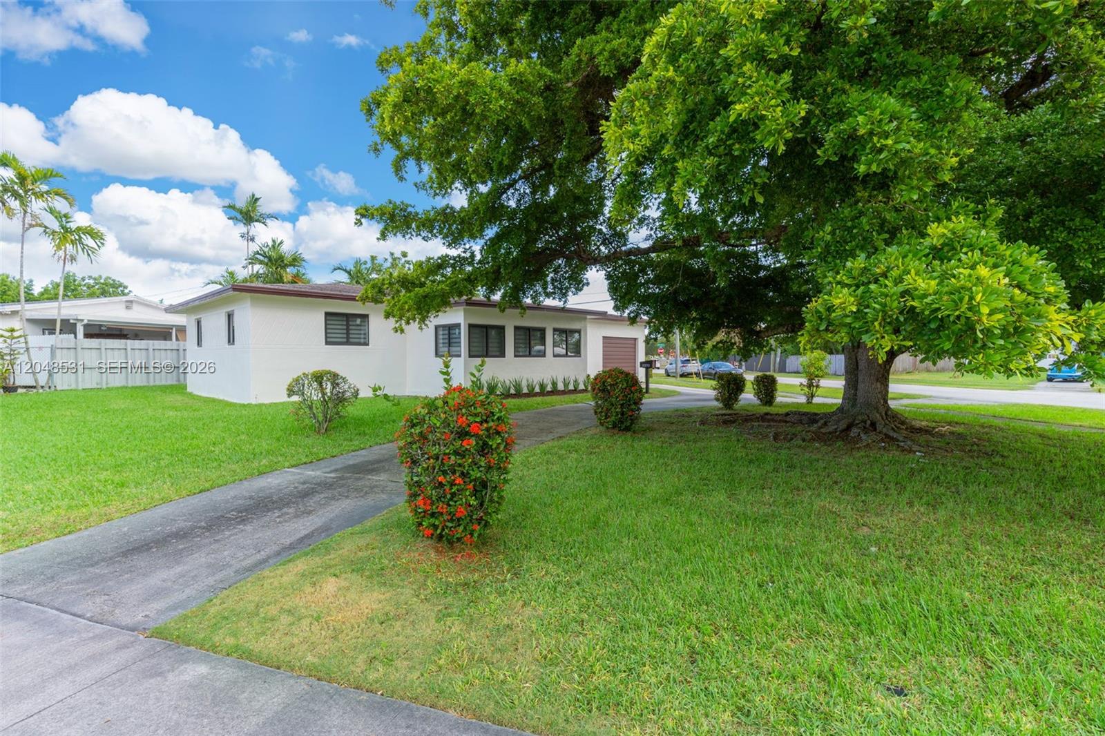 A white single-story house features a large tree, a paved driveway, and manicured landscaping — MIAMI, FL