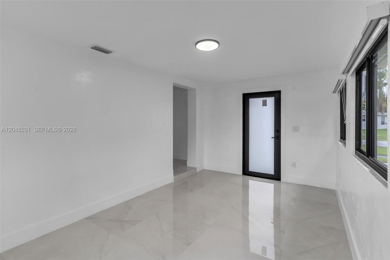 A bright, empty room features white walls, light-colored tile flooring, and a black-framed glass door — MIAMI, FL