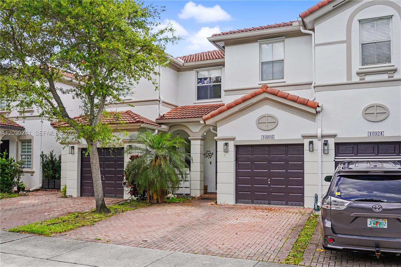 A two-story townhouse exterior features a brick driveway, a brown garage door, and a palm tree — 10918 NW 78th Ter, Doral FL 33178, DORAL, FL