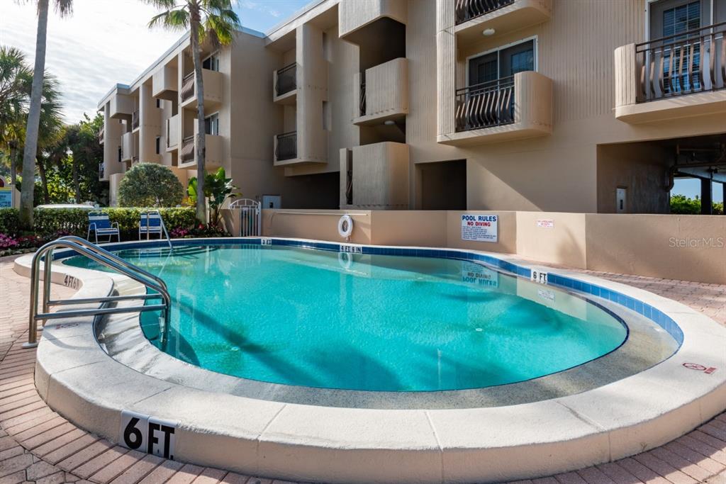 A swimming pool with a curved edge sits in front of a multi-story beige building — 2312 GULF DRIVE N UNIT 103, BRADENTON BEACH, FL