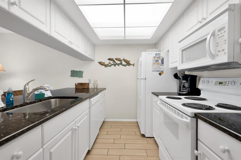 A galley kitchen features white cabinets, black countertops, stainless steel appliances, and a skylight — 2312 GULF DRIVE N UNIT 103, BRADENTON BEACH, FL