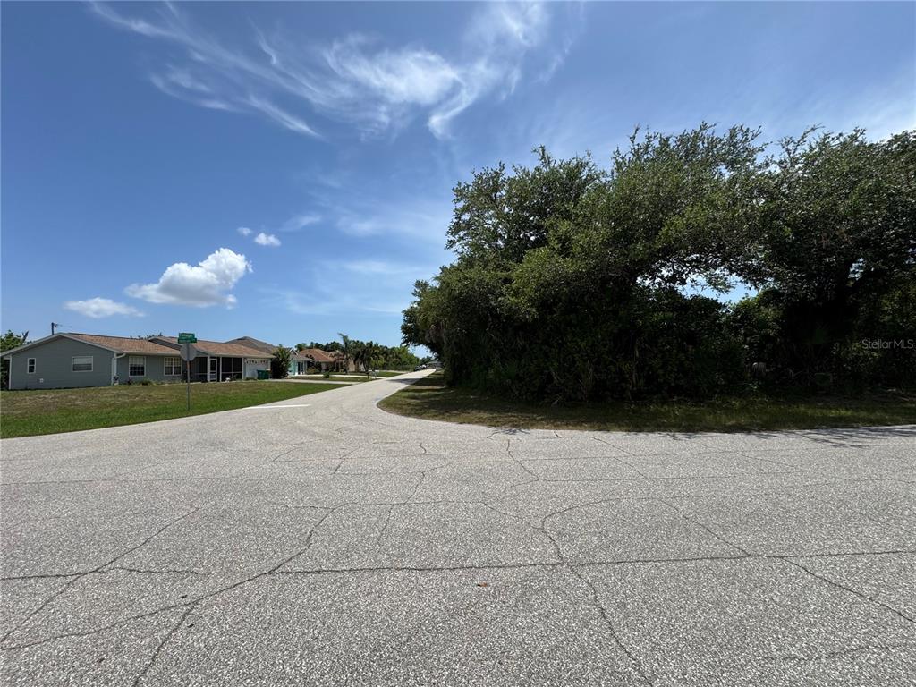 A paved road curves past a residential lot with dense trees and nearby houses — 7251 FINNEGAN STREET, PORT CHARLOTTE, FL