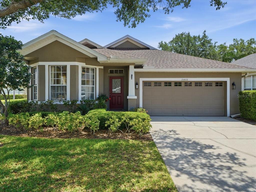 A single-story house with a tan stucco exterior, a red front door, and a two-car garage — 19409 GOLDEN SLIPPER PLACE, LUTZ, FL