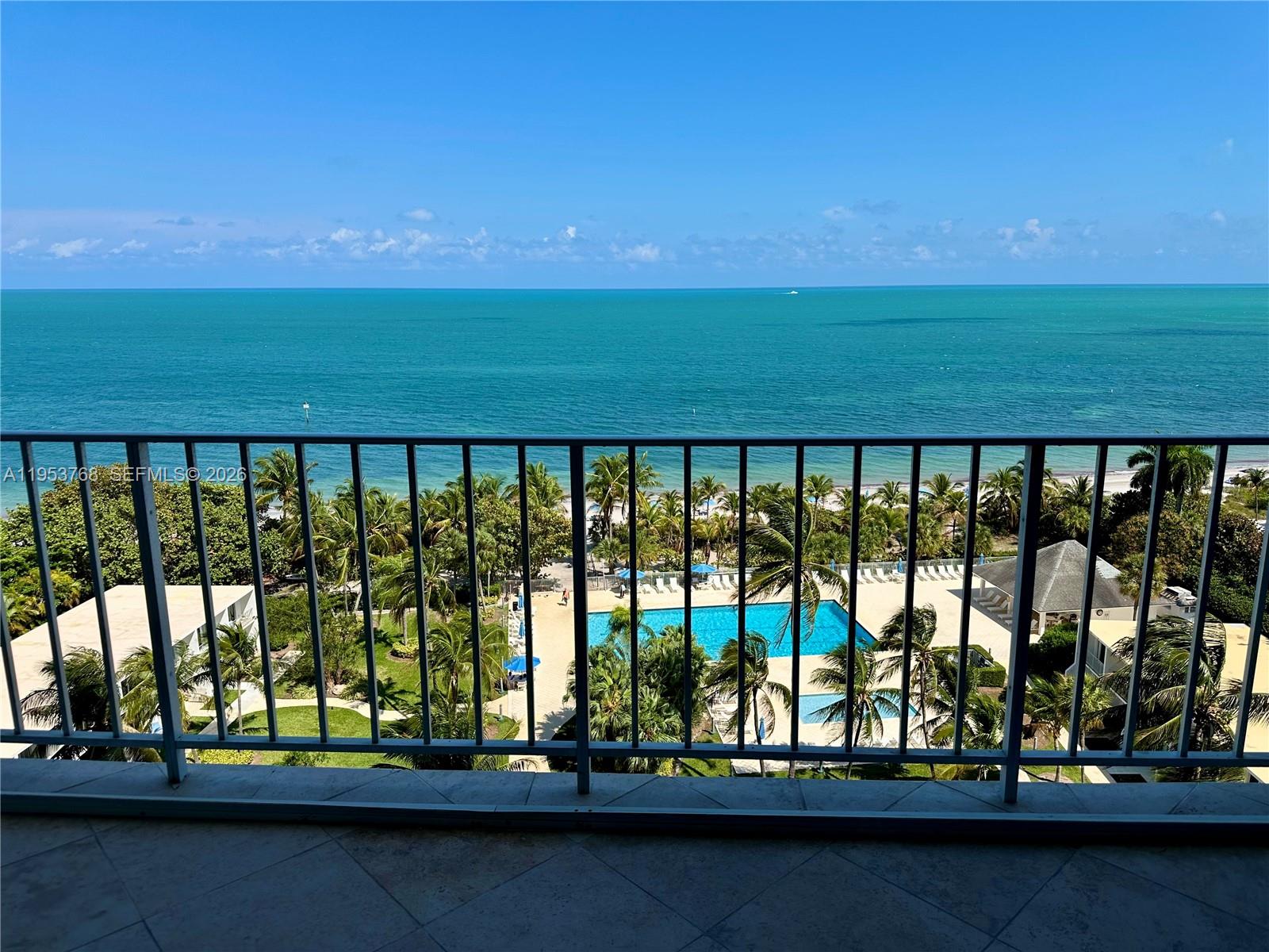 A balcony overlooks a swimming pool, palm trees, and the ocean under a clear sky — 881 Ocean Dr # 10D, Key Biscayne FL 33149, KEY BISCAYNE, FL